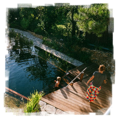Man sieht von schrägt oben auf einen Schwimmteich mit Holzsteg hinunter. Auf dem Steg geht eine junge Frau Richtung Stiege. Im Wasser auf der Stiege steht ein Kind hüfttief im Wasser.